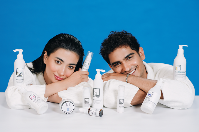 Two Pakistani models in white robes posing with a full range of Dr. Elements skincare and haircare products against a blue background